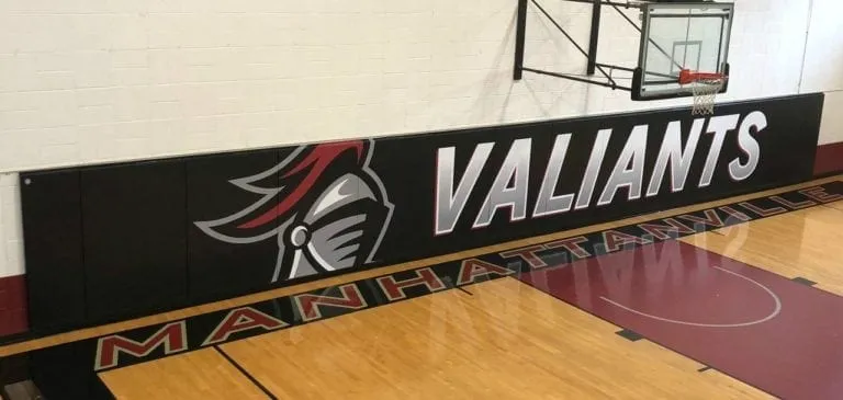 Black gym wall pads printed with the Valiants knight logo above a maple floor with MANHATTAN lettering