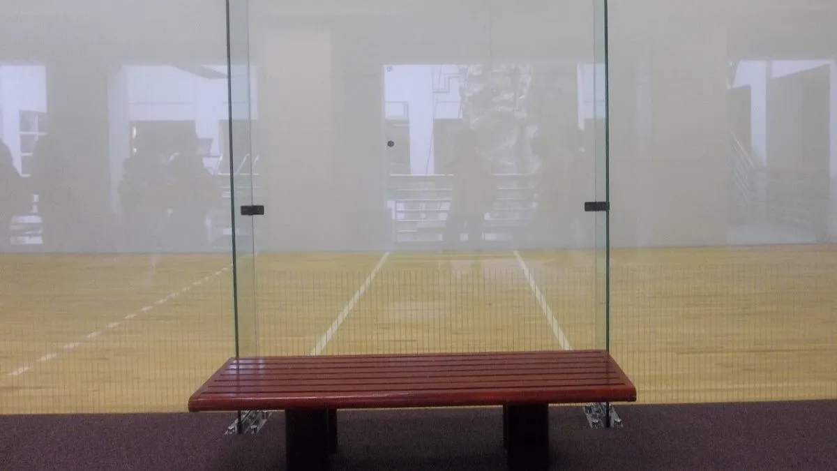 Glass-walled racquetball court with a maple floor and a red bench