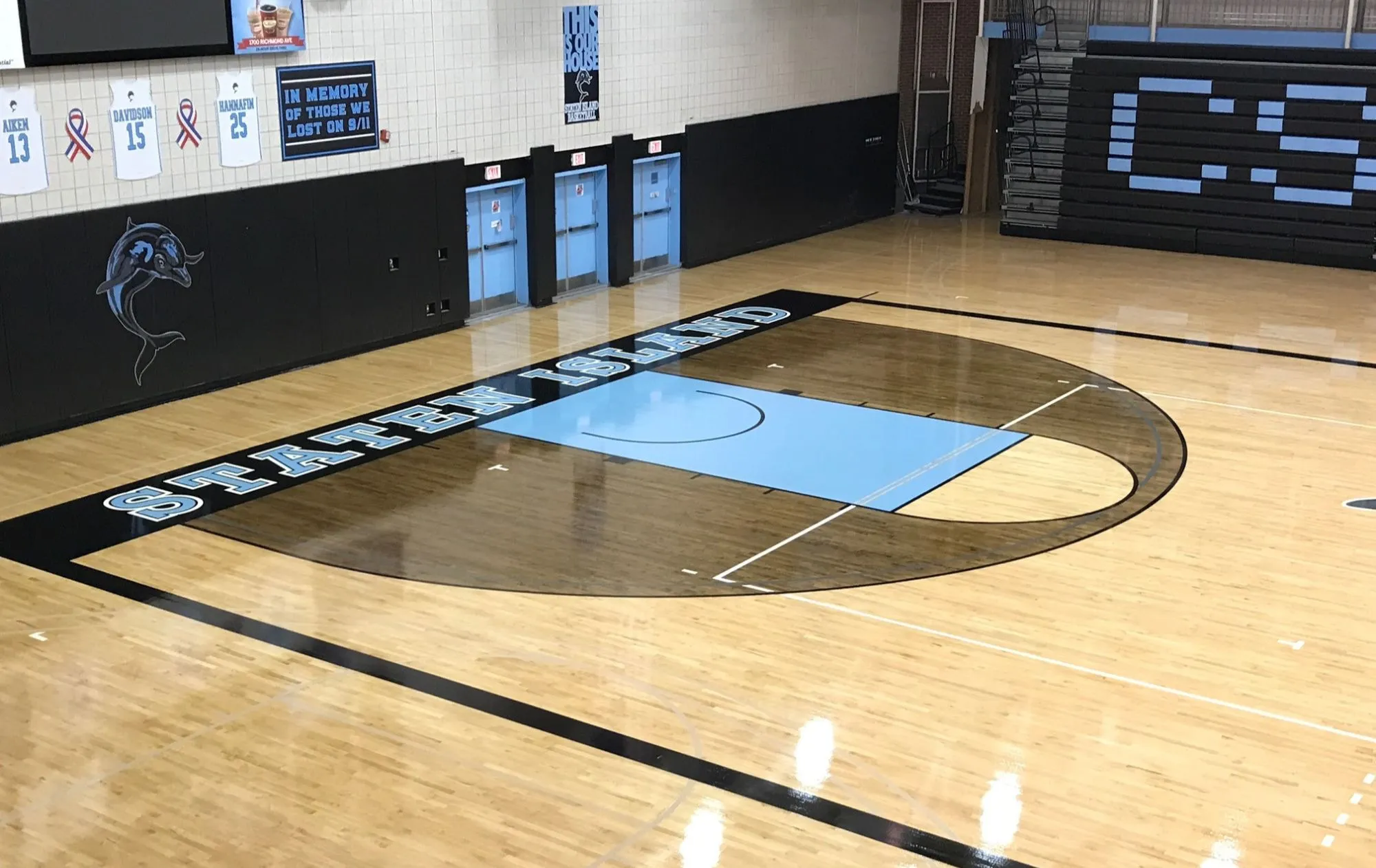 Maple gym floor with a dark-stained three-point area, light blue key and STATEN ISLAND painted on the border