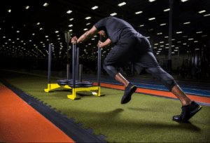 Athlete pushing a weighted sled across green indoor turf in a training gym