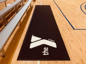 Black courtside runner carpet with a white logo along the sideline of a maple gym floor