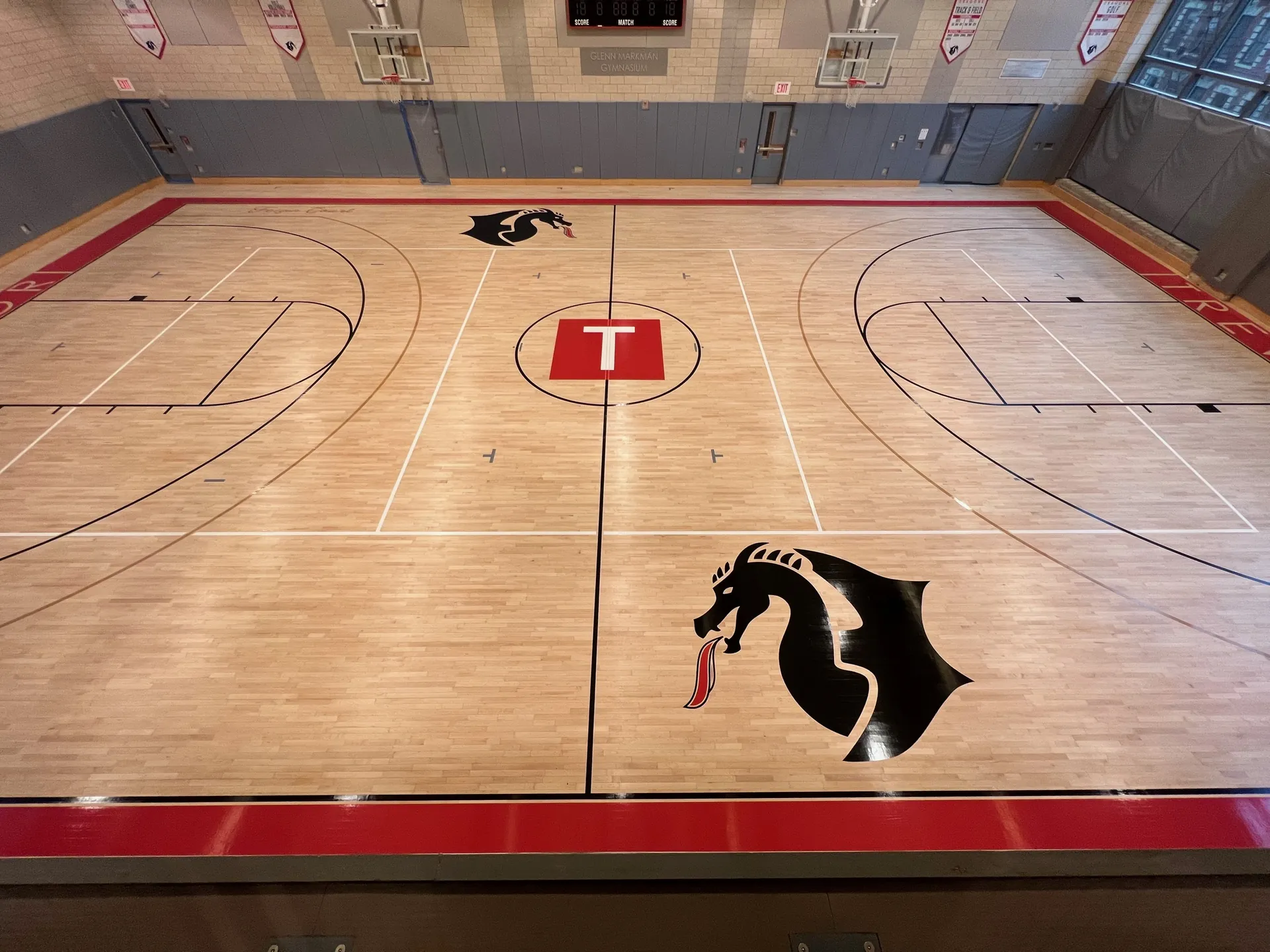 Overhead view of a maple gym floor with a red T center logo, dragon end logos and crisp game lines