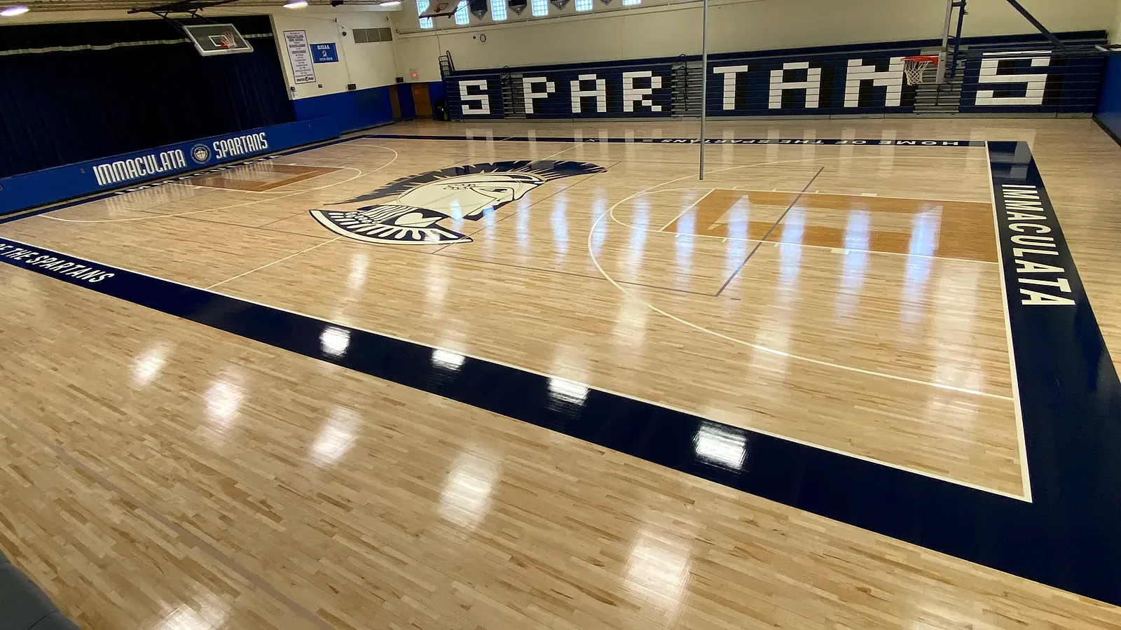 Drone view of the Immaculata Spartans maple gym floor: Spartan center-court logo, navy border with IMMACULATA lettering and a high-gloss finish