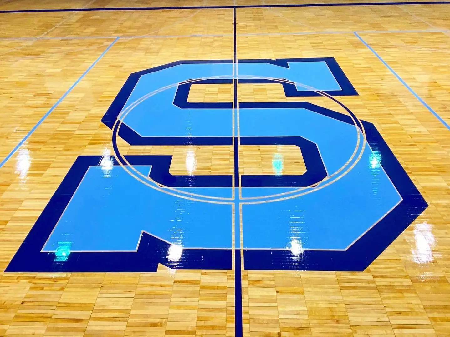 Large light blue S center court logo with navy outline on a maple gym floor
