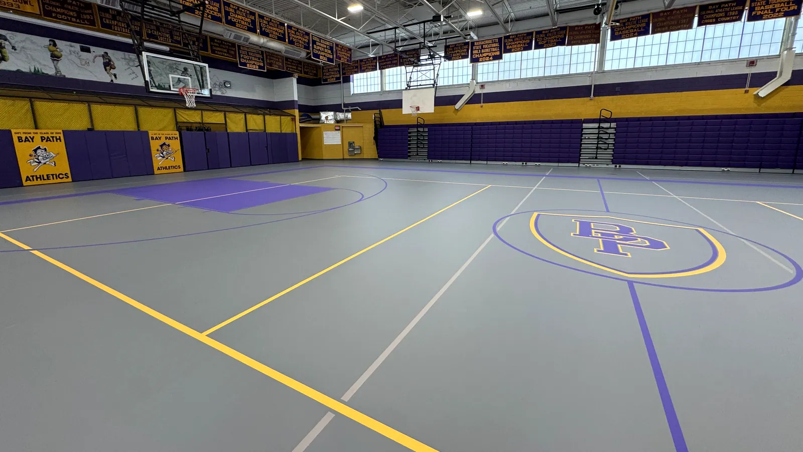 Seamless gray poured gym floor with purple and gold game lines and center-court logo at Bay Path Athletics