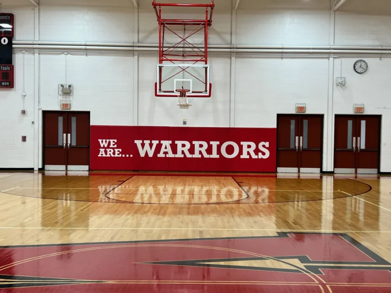 Red printed gym wall pad reading We Are Warriors under a basketball hoop